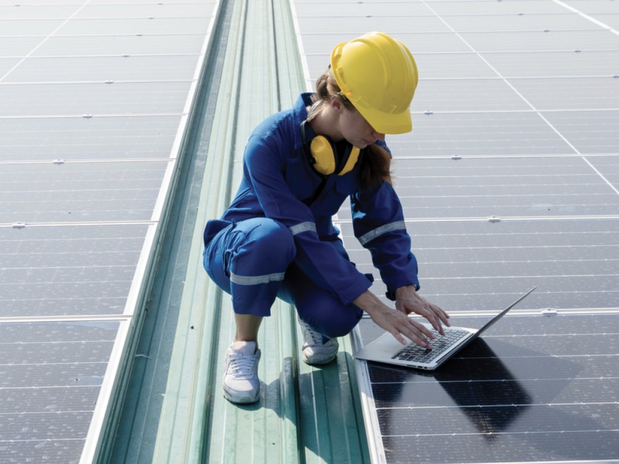 Photograph of woman engineer in yellow hardhat controlling solar panels via laptop